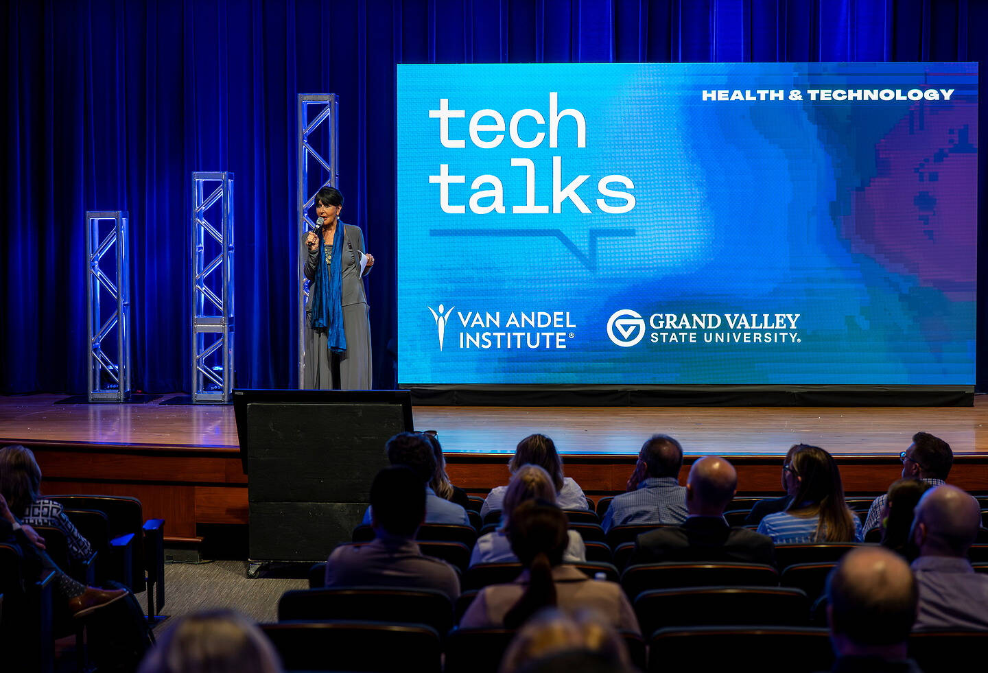 The President stands on stage at a Tech Talks event hosted by the Van Andel Institute and Grand Valley State University, addressing an audience in a dimly lit auditorium.